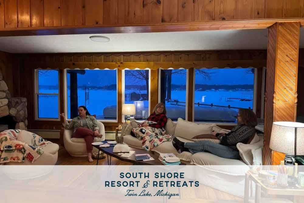 Experience-a-READtreat-at-South-Shore-3 Three people relax in a cozy living room with wooden walls, large windows overlooking a snowy landscape, and a stone fireplace, embodying a peaceful retreat at South Shore Resort and Retreats, ideal for a Lake Michigan getaway.| South Shore Resort & Retreats in West Michigan