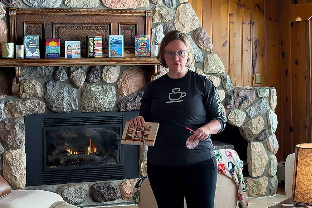 Family-Camp-for-Book-Lovers-1 A person stands in a cozy living room with a stone fireplace, showcasing a book. The warm ambiance of the room highlights the charm of a peaceful retreat, ideal for a relaxing getaway at South Shore Resort and Retreats in West Michigan.| South Shore Resort & Retreats in West Michigan