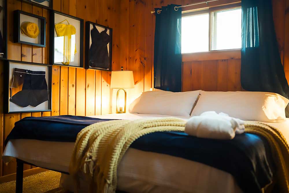 Huron-bedroom-2 A cozy bedroom at South Shore Resort and Retreats features a neatly made bed with white linens, a soft yellow throw, and framed nautical decor on wood-paneled walls, embodying a relaxing getaway in a West Michigan resort.| South Shore Resort & Retreats in West Michigan