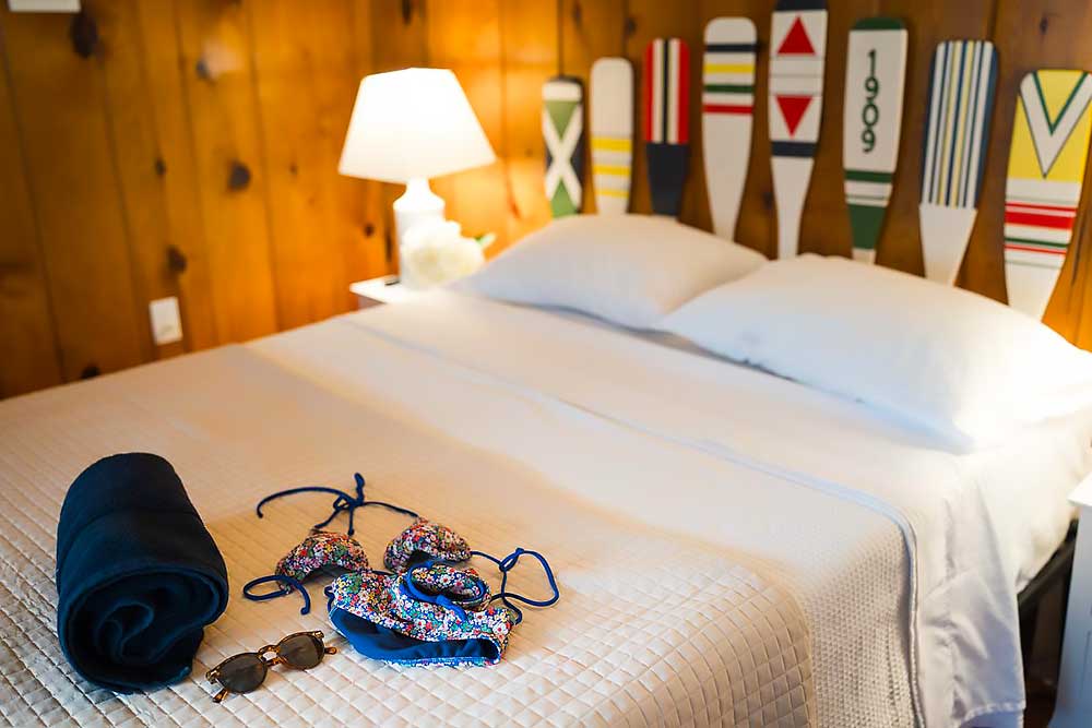 Huron-bedroom-4 Cozy bedroom at South Shore Resort and Retreats with a neatly made bed, wooden paddle decor, a lamp, and a swimsuit and sunglasses on the blanket, ideal for a relaxing getaway in West Michigan.| South Shore Resort & Retreats in West Michigan