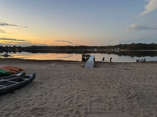 Screenshot-2026-03-25-at-10.41.35-AM A tranquil beach at sunset with a dock extending into calm water, kayaks on the sand, and people enjoying the serene view, perfect for a relaxing getaway at South Shore Resort and Retreats, a beautiful lakefront retreat in West Michigan.| South Shore Resort & Retreats in West Michigan