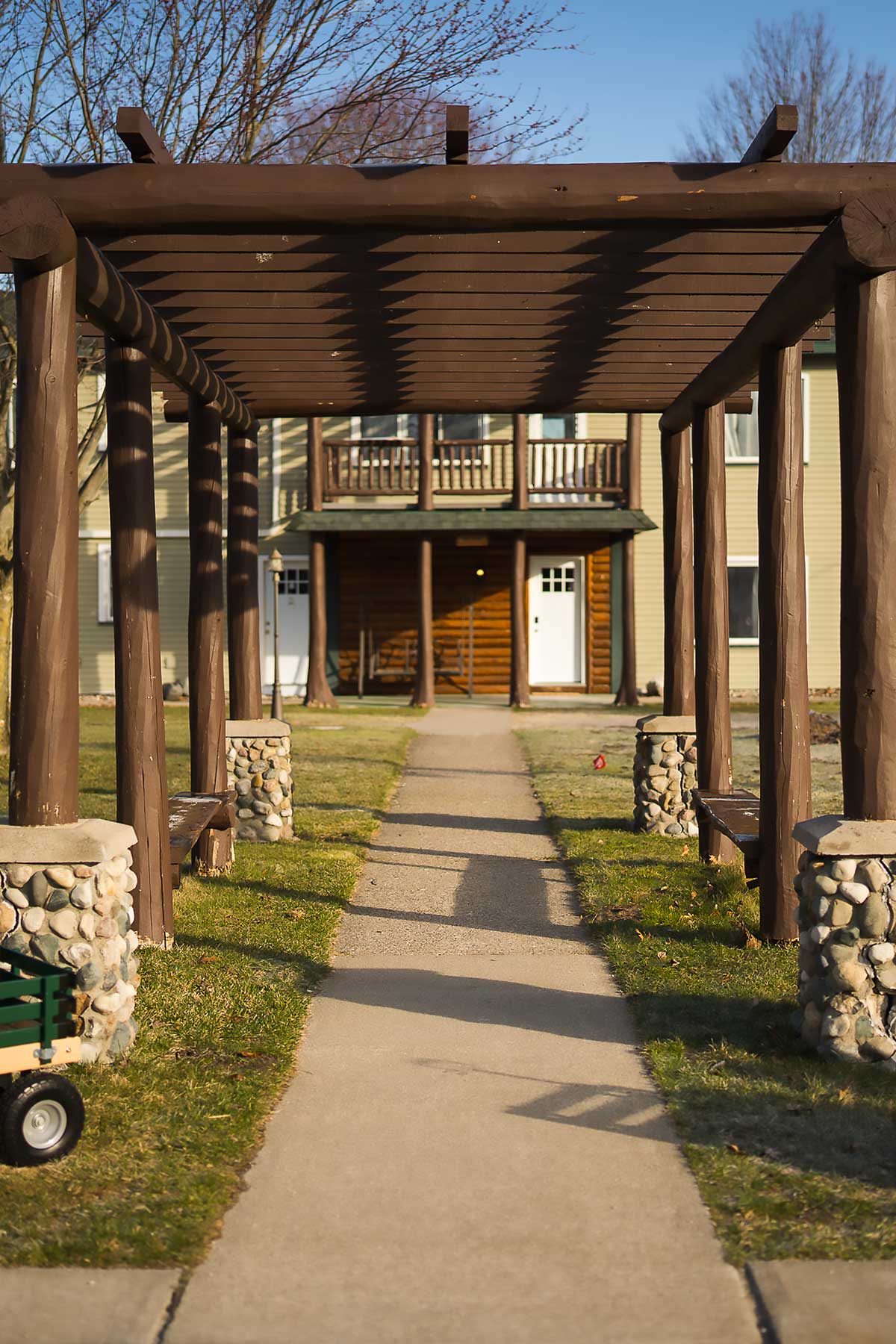 The-lodge-image-1 A wooden pergola with stone pillars lines a sunny path leading to a cozy lodge, ideal for a relaxing getaway at South Shore Resort and Retreats in West Michigan, offering peaceful and scenic lodging.| South Shore Resort & Retreats in West Michigan