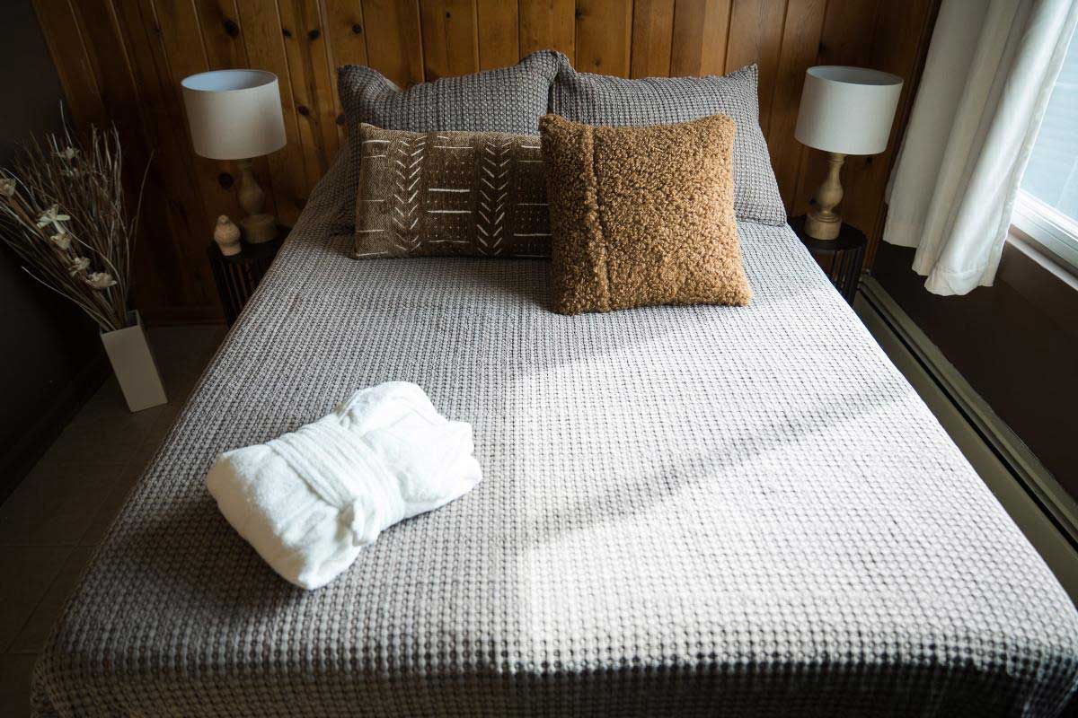 lodge-bedroom-3 A cozy bedroom at South Shore Resort and Retreats features a neatly made bed with textured brown pillows, soft lighting from bedside lamps, and a folded white robe, offering a welcoming and peaceful retreat for guests.| South Shore Resort & Retreats in West Michigan