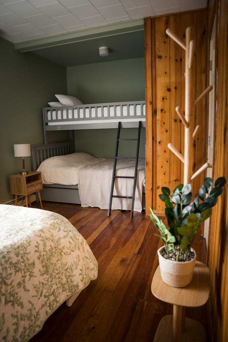 lodge-bedroom-5 A cozy bedroom in a West Michigan vacation rental features a bunk bed with a ladder, wooden floors, a small nightstand with a lamp, and a potted plant, offering a peaceful retreat at South Shore Resort and Retreats.| South Shore Resort & Retreats in West Michigan