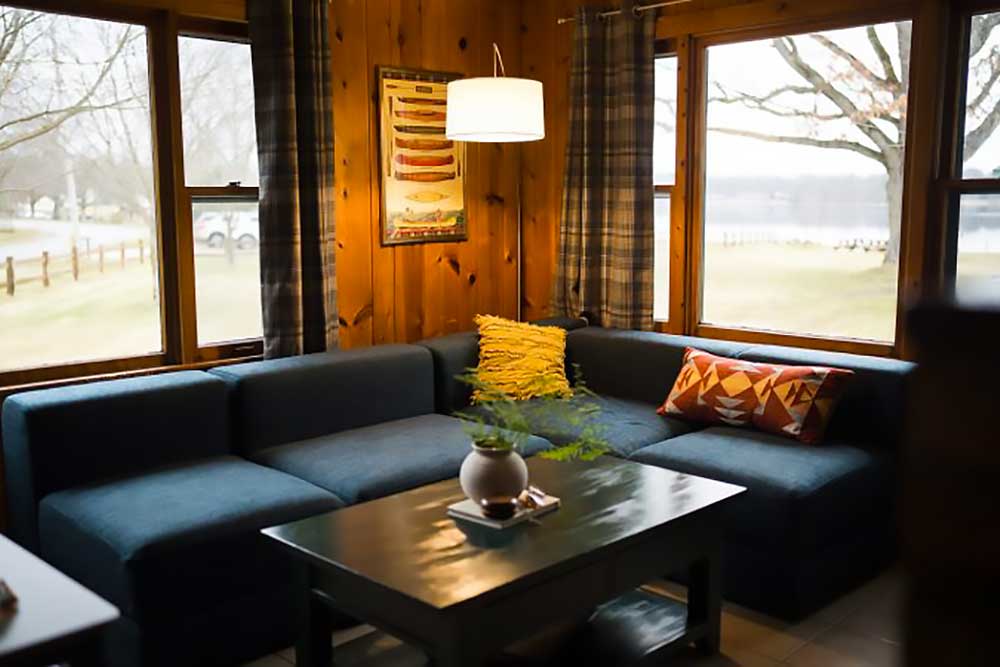 the-huron-image-1 A cozy living room with a blue sectional sofa, colorful pillows, and a wooden table overlooks a serene lake, perfect for a relaxing getaway at South Shore Resort and Retreats in West Michigan.| South Shore Resort & Retreats in West Michigan