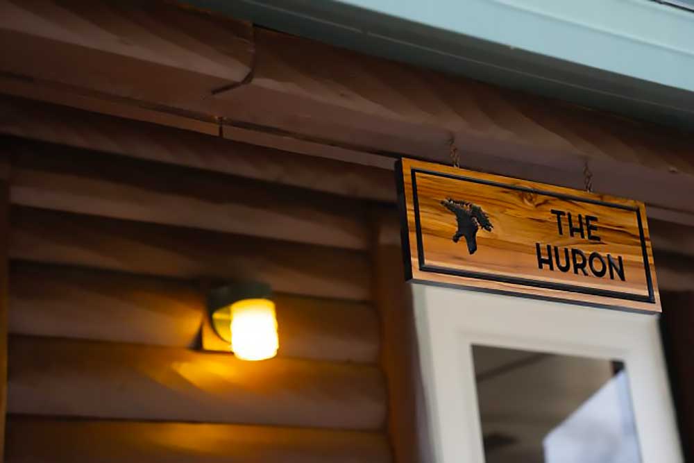 the-huron-image-4 A warmly lit wooden sign reading “The Huron” hangs above a doorway, suggesting a cozy entrance at South Shore Resort and Retreats, ideal for a peaceful retreat in West Michigan.| South Shore Resort & Retreats in West Michigan